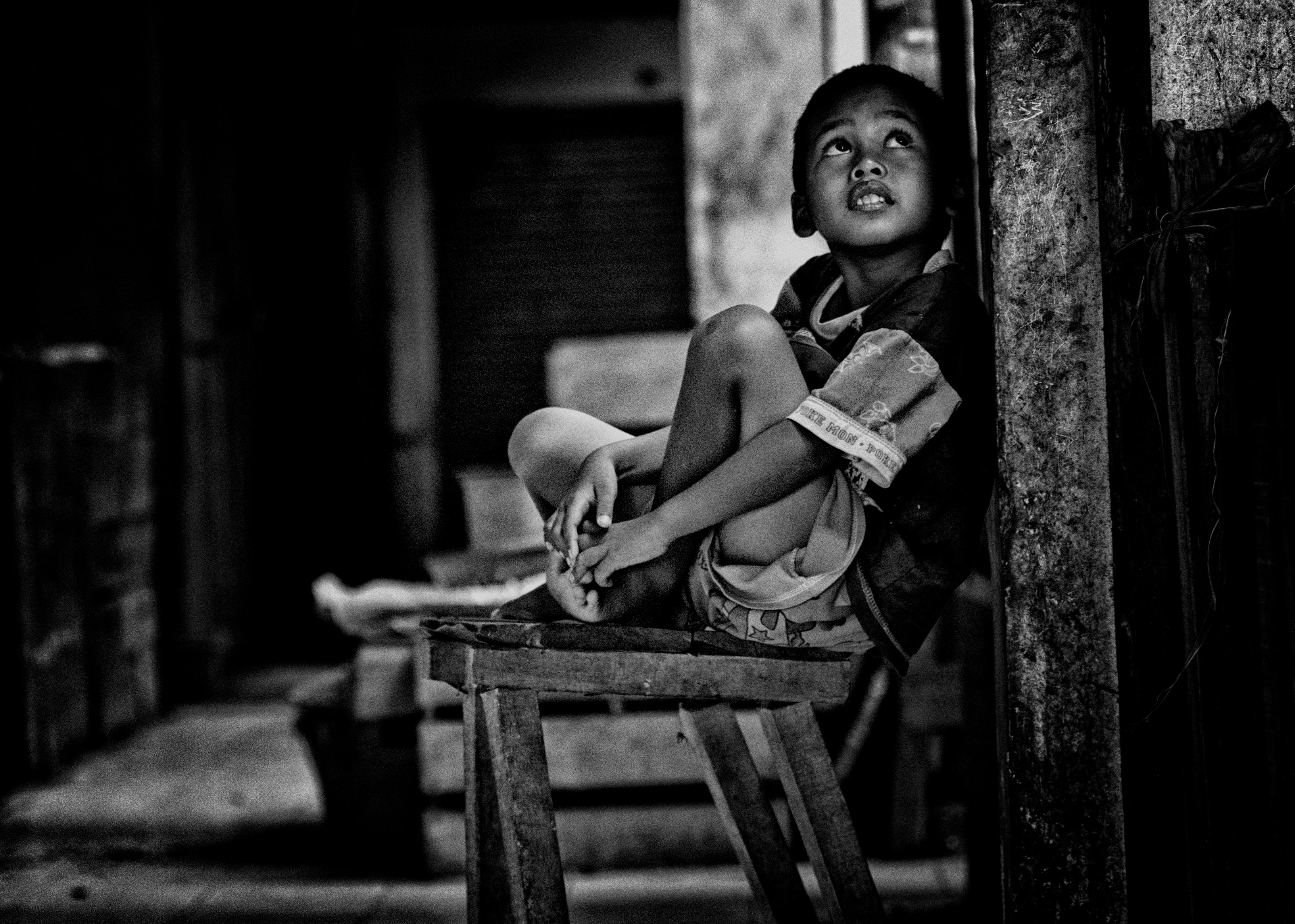 grayscale photo of boy sitting on wooden bench beside wall while looking up